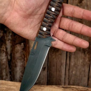 Puede incluir: Un cuchillo de caza negro y marr&oacute;n con un mango de madera texturizado y una hoja negra. El cuchillo tiene un peque&ntilde;o agujero en el mango para un cord&oacute;n.