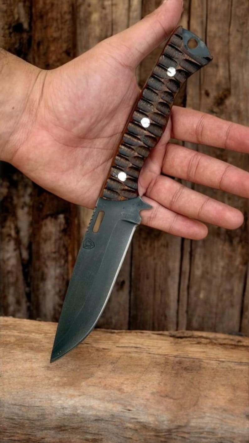 Puede incluir: Un cuchillo de caza negro y marr&oacute;n con un mango de madera texturizado y una hoja negra. El cuchillo tiene un peque&ntilde;o agujero en el mango para un cord&oacute;n.