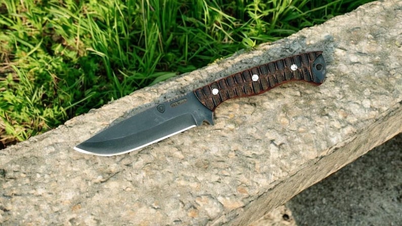 Puede incluir: Un cuchillo de caza negro con mango de madera marr&oacute;n. El cuchillo tiene una hoja negra y un mango negro con un patr&oacute;n de madera marr&oacute;n.