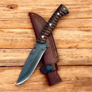 Puede incluir: Un cuchillo de caza negro y marr&oacute;n con una funda de cuero. El cuchillo tiene un mango de madera y una hoja afilada. La funda tiene un dise&ntilde;o decorativo.