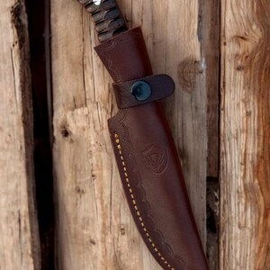 Puede incluir: Una funda de cuchillo de cuero marr&oacute;n con un patr&oacute;n de costura decorativo y un cierre de presi&oacute;n. La funda est&aacute; dise&ntilde;ada para sujetar un cuchillo con un mango de madera.