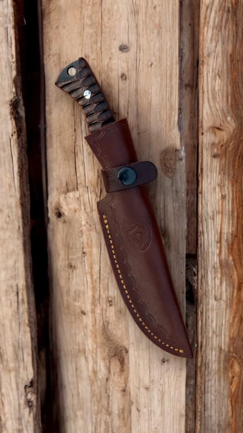 Puede incluir: Una funda de cuchillo de cuero marr&oacute;n con un patr&oacute;n de costura decorativo y un cierre de presi&oacute;n. La funda est&aacute; dise&ntilde;ada para sujetar un cuchillo con un mango de madera.