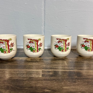 May include: Four white ceramic teacups with a floral design. The cups feature a red, yellow, and green floral pattern with white accents. The cups are arranged in a row on a wooden surface.