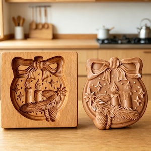 May include: A wooden cookie mold and a baked cookie in the shape of a Christmas ornament. The ornament design includes a bow, candles, stars, pine needles, and pine cones. The mold is made of light-colored wood, and the cookie is brown.