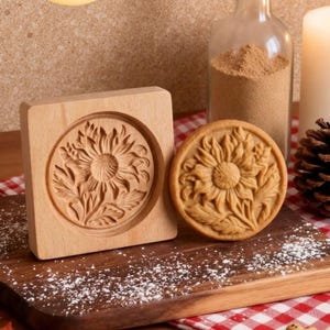 May include: A wooden cookie mold and a baked cookie, both featuring a sunflower design. The mold is square with a circular carved impression. The cookie is round with a detailed sunflower pattern. The items are on a wooden board.