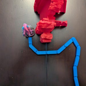 Puede incluir: Configuración de un juego de mesa con una estructura roja texturizada, un camino azul y un pequeño objeto multicolor. Las piezas del juego están dispuestas sobre una superficie marrón oscuro, creando una escena visualmente atractiva.
