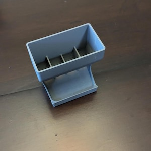 May include: A gray plastic organizer with a rectangular top section divided into three compartments. The organizer has a curved base and is designed to hold small items. The object is made of a solid, smooth material.