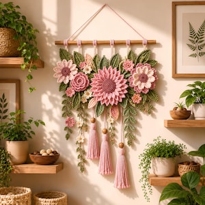 Può includere: Una decorazione da parete con fiori rosa e foglie verdi realizzati all'uncinetto, fissati a un bastone di legno orizzontale. La composizione include tre nappe di filo rosa appese a cordini con perline di legno.