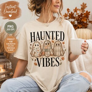 Op de afbeelding: Een crèmekleurig t-shirt met een afbeelding van drie geesten met verschillende patronen, waaronder bloemen, patchwork en ruiten. De tekst "HAUNTED VIBES" is in het zwart boven en onder de geesten gedrukt, omringd door kleine herfstblaadjes en eikels.
