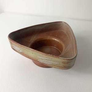 Könnte beinhalten: Eine handgefertigte Keramikschale mit dreieckiger Form und Sockel. Die Schale hat eine warme, erdige Farbpalette mit Braun- und Beigetönen. Der Rand hat ein helleres, strukturiertes Design.