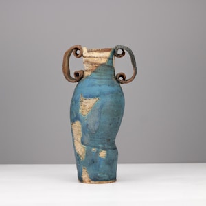 Può includere: Un vaso in ceramica alto con due manici prominenti e arricciati. Il corpo presenta una finitura ruvida e screziata con toni blu intenso e beige terroso. La superficie ha un aspetto grezzo e organico con macchie di argilla visibili.