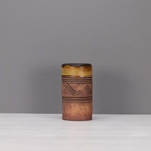 May include: A cylindrical ceramic vase with a textured brown body featuring horizontal grooved lines and a zigzag pattern. The upper section of the vase has a smooth, contrasting mustard-yellow glaze that extends to the top rim of the vessel.