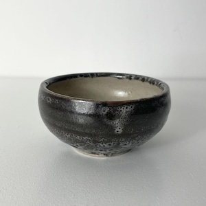May include: A small, handmade ceramic bowl. The exterior is dark and textured, while the interior is light-coloured. The bowl has a rounded shape and an uneven rim, highlighting its handcrafted quality. The exterior has a speckled pattern and a glossy finish.