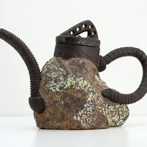 May include: A unique ceramic teapot with a textured, rock-like body in shades of brown and green. It features a dark, industrial-style handle, spout, and lid with circular cutouts. The teapot has a distinctive, artistic design.