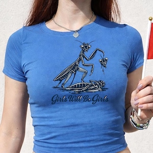 Könnte beinhalten: Blaues T-Shirt mit einer Grafik einer Gottesanbeterin, die ein Herz über einem toten Insekt hält. Der Text "Girls Will Be Girls" ist darunter gedruckt. Eine Person hält eine kleine Flagge.