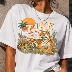 May include: A white t-shirt featuring a graphic print of a tiger resting in a field of flowers and palm trees. Above the tiger, the text reads "TAKE IT EASY" in a retro-style font, set against a large orange sun. The design includes green foliage.