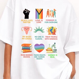 May include: White t-shirt featuring various colorful graphics and text. Designs include a raised fist, a rainbow-colored 'Love is Love' symbol, and the words 'Black Lives Matter'. Other graphics include a uterus, books, and a rainbow heart.