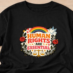 Op de afbeelding: Zwart T-shirt met een kleurrijke afbeelding met de tekst "HUMAN RIGHTS ARE ESSENTIAL". Het ontwerp bevat een regenboog, een duif, rozen, sterren en een weegschaal. Het T-shirt is gemaakt van zacht materiaal.