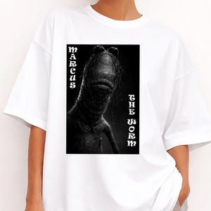 May include: White t-shirt with a black and white graphic of a creature. The text "MARCUS THE WORM" is vertically aligned on either side of the image. The graphic is centered on the front of the shirt.