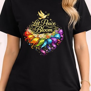 Puede incluir: Camiseta negra con un diseño floral colorido y el texto "Let Peace Bloom". El diseño incluye una cinta arcoíris, flores y una paloma dorada. La camiseta es de manga corta.