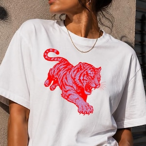 Può includere: T-shirt bianco con un disegno di tigre rossa e rosa. La tigre è raffigurata in una posa dinamica, con la bocca aperta e gli artigli estesi. La maglietta ha maniche corte e girocollo.
