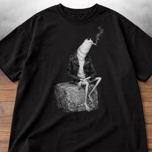 Può includere: T-shirt nero con un'illustrazione in scala di grigi di una creatura con giacca di pelle e occhiali da sole, che fuma una sigaretta. La creatura è seduta su una pietra, con un design unico.