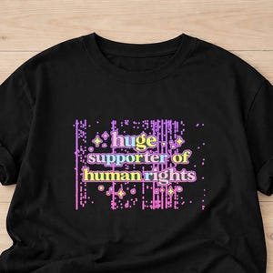 Op de afbeelding: Zwart T-shirt met de tekst "huge supporter of human rights" in een pixelachtige, veelkleurige letter. Het ontwerp bevat paarse, roze, gele en blauwe tinten. Het T-shirt is gemaakt van zachte stof.