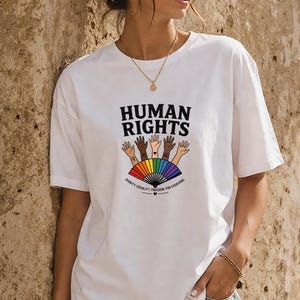 Op de afbeelding: Wit T-shirt met de zwarte tekst "HUMAN RIGHTS" boven een afbeelding van open handen in verschillende huidtinten en een regenboogwaaier. De tekst "DIGNITY. EQUALITY. FREEDOM. FOR EVERYONE." staat eronder.