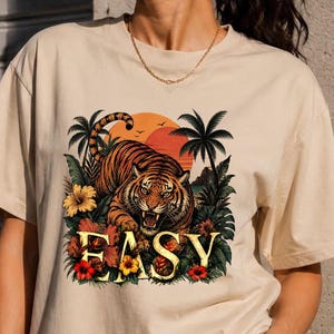 Può includere: T-shirt beige con un'illustrazione di tigre e la scritta "EASY" in oro. La tigre è arancione e nera, sullo sfondo di un tramonto con palme e fiori. Una t-shirt casual e confortevole.