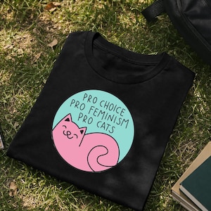 Op de afbeelding: Zwart T-shirt met een ronde afbeelding met een roze kat en de woorden "Pro Choice, Pro Feminism, Pro Cats" in witte tekst op een lichtblauwe achtergrond. Het shirt ligt op groen gras.