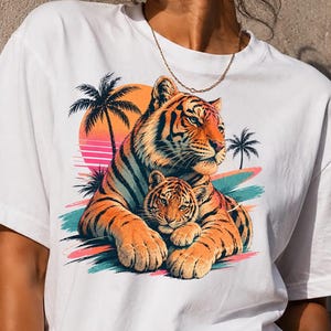 Può includere: Una t-shirt bianca a maniche corte con una stampa grafica raffigurante una tigre adulta e un cucciolo che riposano insieme. Il design include un vivace tramonto arancione, sagome di palme e tocchi di pennellate color ottanio e rosa.