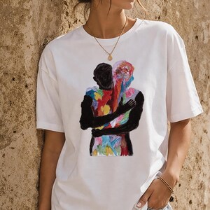 Op de afbeelding: Wit T-shirt met een kleurrijk abstract schilderij van twee figuren die elkaar omarmen. Het kunstwerk maakt gebruik van een levendig palet van rood, blauw, geel en groen. Het shirt is een klassieke ronde hals.