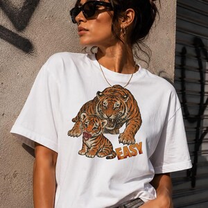 Può includere: T-shirt bianca con grafica di tigre e cucciolo nei toni dell'arancione e del marrone. La parola "EASY" è stampata in arancione. La maglietta ha maniche corte e girocollo.