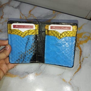 May include: A bi-fold wallet featuring a black exterior and an interior with two card slots. The slots have a textured blue and gold snakeskin-like pattern. Each slot contains a white card with the text Genuine Leather printed in red.
