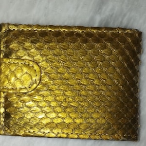 May include: A rectangular wallet featuring a metallic gold finish and a textured snakeskin pattern. The wallet includes a matching gold-colored tab closure on the left side, secured with visible stitching along the edges of the entire accessory.