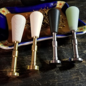 May include: Four tobacco pipe tampers with different handle colors: light wood, white, black, and green. Each tamper has a metal stem and a flat, circular base. The tampers are arranged in a row on a dark wooden surface.
