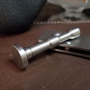 May include: A silver-colored, cylindrical bottle stopper with a flat, circular top. The stopper is lying on a closed, silver-colored knife, set against a brown leather surface. A black object is in the background.