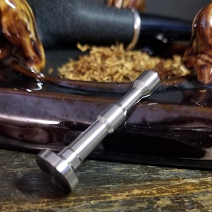 May include: A close-up of a shiny, silver metal pipe tool. The tool has a cylindrical body with a flat, rounded end and is set against a backdrop of a pipe, tobacco, and a decorative tray.