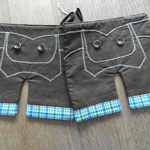 Könnte beinhalten: Zwei braune Ofenhandschuhe in Form von Lederhosen-Shorts. Jeder Handschuh hat ein Taschen-Design mit zwei Knöpfen und weißen Nähten. Die Manschetten sind blau und weiß kariert. Eine weiße Schnur ist zum Aufhängen befestigt.