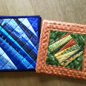 Könnte beinhalten: Zwei gesteppte Topflappen. Einer ist hauptsächlich blau mit einem diagonalen Muster, der andere orange mit einem grünen Zentrum und einer Schlaufe zum Aufhängen. Die Topflappen sind quadratisch und handgefertigt.