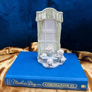 May include: A porcelain figurine of a window with curtains and two white dogs. The window has a small plant on the sill and a table with a book on it. The figurine is sitting on a blue book with the title "Mother's Day on Coronation St."
