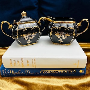 May include: Black and gold ceramic creamer and sugar set with floral designs. The set is on top of two books.