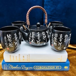 May include: A black ceramic tea set with a brown handle on the teapot and four cups. The cups and teapot are decorated with white calligraphy.