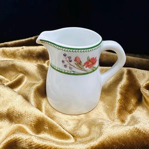 May include: A white ceramic creamer with a floral design in shades of pink, green, and purple. The creamer has a rounded body and a small handle.