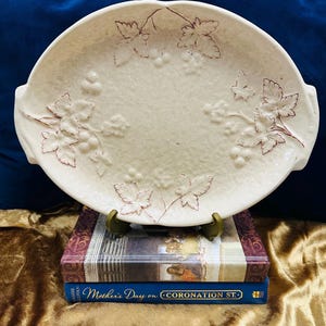 May include: Cream-coloured, oval serving platter with an embossed grape and leaf pattern. The platter has two handles and is displayed on a stack of books. The books are titled "Mother's Day on Coronation St."