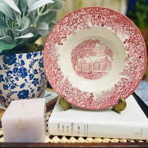 Può includere: Una zuppiera in ceramica rosa e bianca con un motivo floreale e una scena di una casa con alberi. La zuppiera è su un supporto in metallo dorato e un libro bianco con lettere dorate.