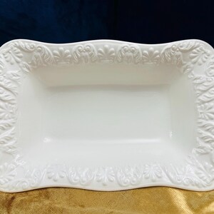 Puede incluir: Plato de servir de cerámica blanca con un diseño ornamentado y en relieve alrededor del borde. El plato es rectangular con esquinas redondeadas y un borde ligeramente ondulado.