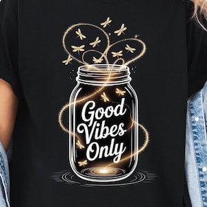 Może przedstawiać: Czarny t-shirt z grafiką szklanego słoika wypełnionego świecącym światłem i latającymi ważkami. Napis Good Vibes Only jest nadrukowany na biało na środku słoika, otoczony wirującymi smugami złotego światła.