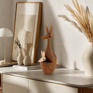 May include: A wooden kangaroo figurine with a joey in its pouch. The figurine is brown and has a natural wood finish. It is sitting on a white countertop in front of a white wall.