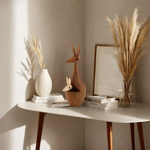 May include: A wooden kangaroo sculpture with a joey in its pouch. The sculpture is brown and has a natural wood finish. It is sitting on a white table with a vase and books.
